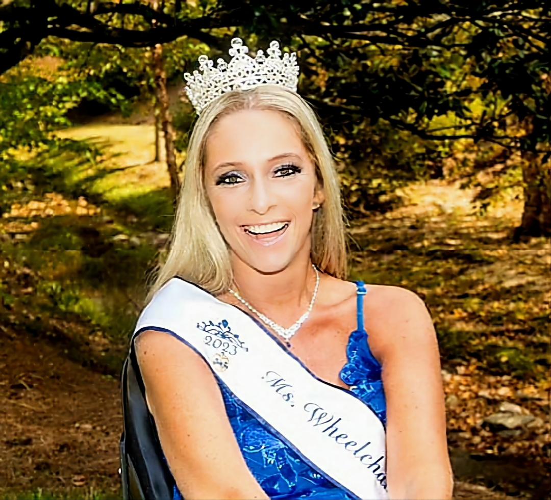 Pictured is a Caucasian female with straight blonde hair past her shoulders. She is wearing a blue colored top with thin straps and a sash across her body that reads 2023 Ms. Wheelchair America. She is in the seated position.